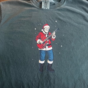 May include: Dark green Comfort Colors t-shirt featuring a graphic of Santa Claus playing a red electric guitar. Santa is wearing a red suit, blue pants, and black boots. White snowflakes are scattered around the image.