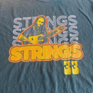 Op de afbeelding: Blauw-grijs T-shirt met een afbeelding van een persoon die gitaar speelt, en het woord "STRINGS" in oranje en wit. Het nummer "33" is geel gedrukt onder het woord "STRINGS".