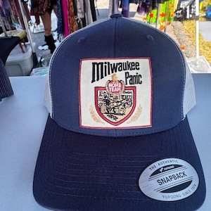 May include: Navy blue trucker hat with a white mesh back and a curved brim. The front features a patch with the words "Milwaukee Panic" and a graphic. The hat has a snapback closure.