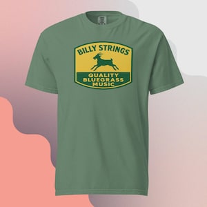 Könnte beinhalten: Ein grünes T-Shirt mit einem Logo mit der Aufschrift "Billy Strings" über einer Ziegensilhouette und den Worten "Quality Bluegrass Music". Das Logo ist gelb und grün. Das Shirt hat eine einfarbige grüne Farbe.