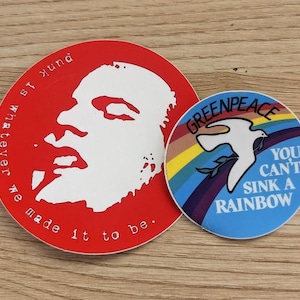 Może przedstawiać: Dwie okrągłe naklejki. Jedna czerwona naklejka przedstawia biały profil i tekst "punk is whatever we made it to be". Druga naklejka jest niebieska z tęczą, gołębiem i tekstem "Greenpeace You Can't Sink a Rainbow."