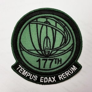 May include: A green and black embroidered patch with a stylized design of a globe and a plane. The text "177th" and "TEMPUS EDAX RERUM" are embroidered in white on the patch.