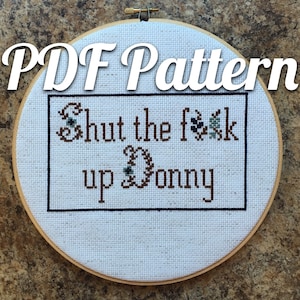 May include: A cross-stitch embroidery hoop with the text "PDF Pattern" at the top. The cross-stitch design says "Shut the f**k up Donny" in brown thread with decorative floral accents. The embroidery is framed in a black border.