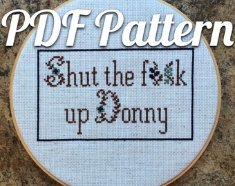 May include: A cross-stitch embroidery hoop with the text "PDF Pattern" at the top. The cross-stitch design says "Shut the f**k up Donny" in brown thread with decorative floral accents. The embroidery is framed in a black border.