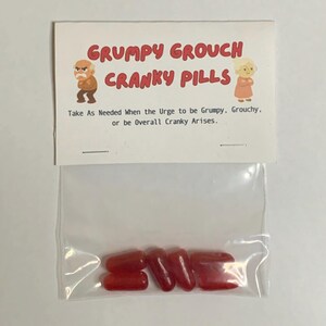 May include: A clear plastic bag containing several red, oblong-shaped candies. The bag is attached to a card with the text "GRUMPY GROUCH CRANKY PILLS" and instructions. The card features cartoon illustrations of an elderly man and woman.