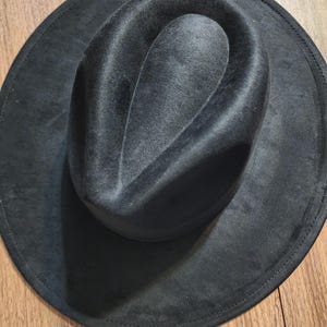 May include: A dark gray fedora hat with a wide brim. The hat has a soft, velvety texture and a classic teardrop crown. The brim is stitched around the edge. The hat is sitting on a wooden surface.