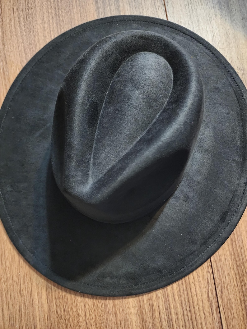 May include: A dark gray fedora hat with a wide brim. The hat has a soft, velvety texture and a classic teardrop crown. The brim is stitched around the edge. The hat is sitting on a wooden surface.