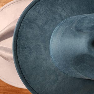 May include: Two fedora hats in a close-up shot. One is a light gray color, and the other is a teal color. The hats are made of a soft, velvety material. The hats are sitting on a wooden surface.