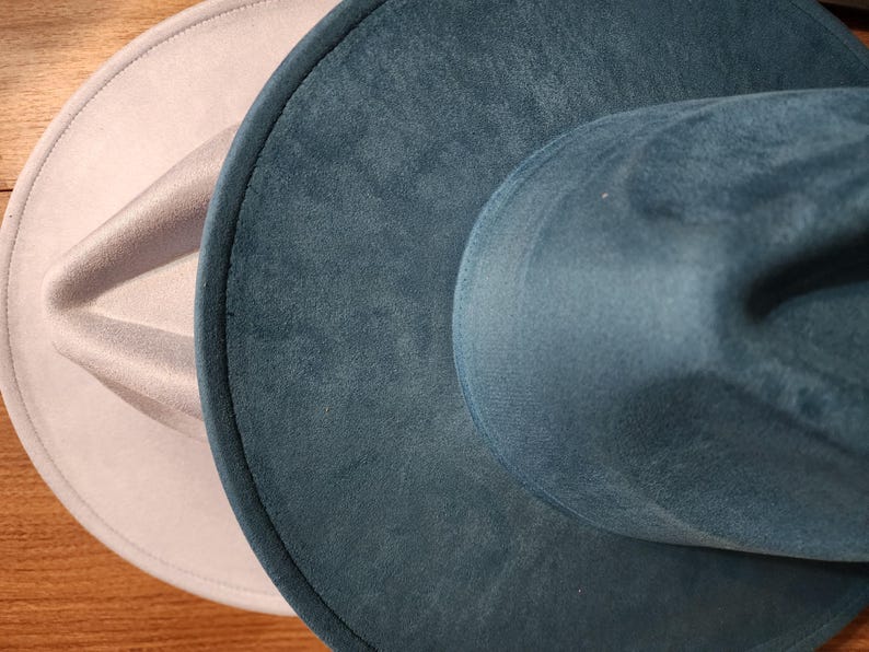 May include: Two fedora hats in a close-up shot. One is a light gray color, and the other is a teal color. The hats are made of a soft, velvety material. The hats are sitting on a wooden surface.