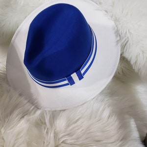 May include: A blue fedora hat with a white brim and blue stripes. The hat is sitting on a white fluffy surface.