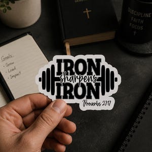 May include: A white sticker with black text that reads "IRON sharpens IRON Proverbs 27:17" in a barbell shape. The sticker is held in a hand, with a black notebook, a Bible, and a mug in the background.