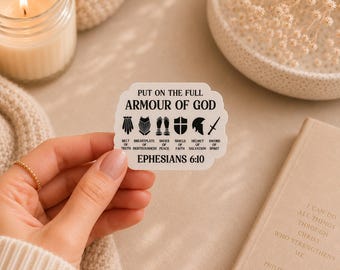 May include: A white sticker with the text "PUT ON THE FULL ARMOUR OF GOD" and illustrations of armor. The sticker also includes the text "EPHESIANS 6:10". A hand holds the sticker near a lit candle and a book.