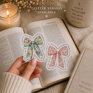 May include: Two bow-shaped stickers with floral designs are held over an open book. One bow is blue and green, the other is pink. A lit candle, a mug with text, and a closed book are also visible. Text on the stickers reads: "She is clothed with strength and dignity."