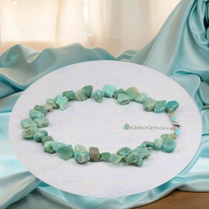 May include: A light green gemstone necklace made of irregularly shaped stones. The necklace is on a white surface and has a silver clasp.