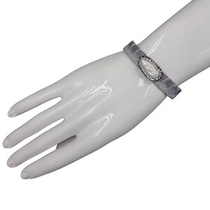 May include: A gray velvet bracelet with a silver and white oval-shaped charm. The charm is decorated with small clear rhinestones.