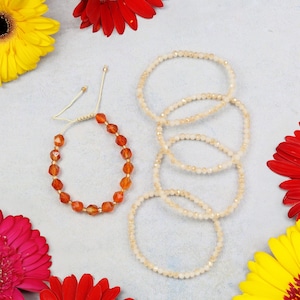 May include: Two styles of beaded bracelets are shown. One is burnt orange with a macrame closure. The other is a set of four light beige bracelets. The bracelets are on a light blue surface, decorated with red and yellow flowers.