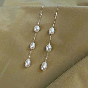 Puede incluir: Elegantes pendientes colgantes con perlas ovaladas y brillantes suspendidas de delicadas cadenas de plata. Cada pendiente presenta tres perlas, creando un diseño elegante y alargado.