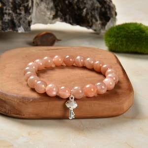 May include: A peach-colored sunstone beaded bracelet with a silver key charm. The bracelet is on a wooden surface.