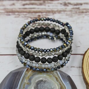 Puede incluir: Un brazalete de m&uacute;ltiples capas con cuentas de lava negra y cuentas de vidrio iridiscente. El brazalete est&aacute; apilado con m&uacute;ltiples hileras de cuentas, creando un aspecto en capas y texturizado.