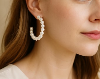 May include: A close-up of a person wearing a pair of white pearl hoop earrings. The earrings are made of small, round pearls and are in a classic hoop shape. The person is wearing a cream-colored top.