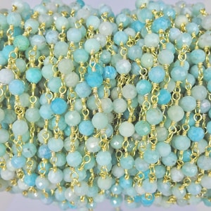 Puede incluir: Primer plano de un hilo de cuentas de piedras preciosas en tonos azules y verdes. Las cuentas redondas y facetadas están ensartadas en cadenas doradas. Las cuentas tienen aproximadamente 0,5 cm de diámetro. El efecto general es delicado y elegante.