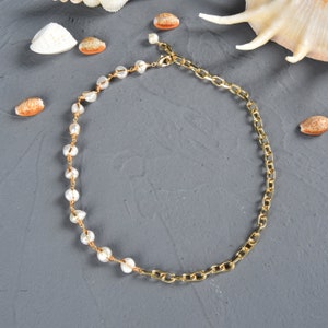 May include: A gold chain necklace with a pearl strand on one side. The necklace is on a gray background with seashells.