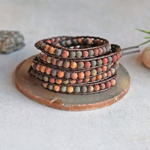 May include: A multi-strand bracelet made with brown leather cord and colorful jasper beads. The bracelet is wrapped around a light gray stone coaster.