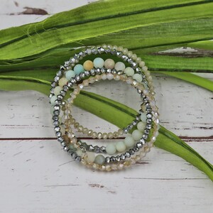 Puede incluir: Un conjunto de cinco pulseras con una mezcla de cuentas verde claro y beige, y cuentas plateadas. Las pulseras est&aacute;n superpuestas.