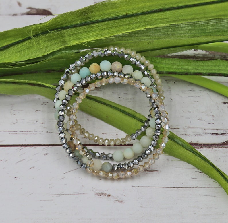 Puede incluir: Un conjunto de cinco pulseras con una mezcla de cuentas verde claro y beige, y cuentas plateadas. Las pulseras est&aacute;n superpuestas.
