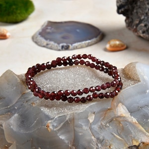 May include: A double strand bracelet made of small, dark red garnet beads. The bracelet is on a light gray, textured surface.