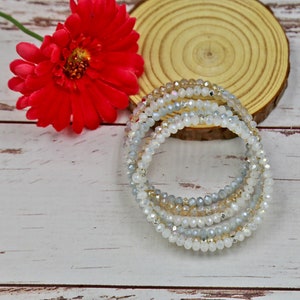 Puede incluir: Un conjunto de cuatro pulseras de cuentas en tonos blanco, gris y beige. Las pulseras est&aacute;n hechas de cuentas peque&ntilde;as y redondas y est&aacute;n apiladas unas sobre otras.