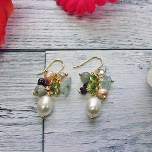 Könnte beinhalten: Ein Paar goldfarbene Ohrringe mit einer Ansammlung bunter Perlen und einer großen weißen Perle. Die Ohrringe haben eine Mischung aus grünen, blauen, rosa und grauen Perlen, mit einer tropfenförmigen Perle unten.