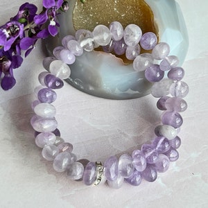 May include: A purple amethyst gemstone bracelet with a silver charm. The bracelet is made of smooth, rounded beads.
