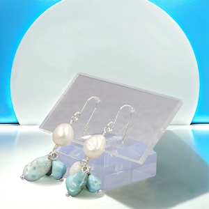 May include: A pair of dangle earrings featuring white freshwater pearls and light blue larimar stones. The earrings have silver-colored hooks and are displayed on a clear acrylic stand against a blue and white background.