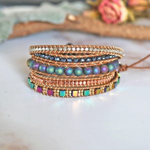 May include: A multi-layered bracelet with brown leather cord, iridescent beads, and square-shaped beads. The bracelet has a silver and copper finish.