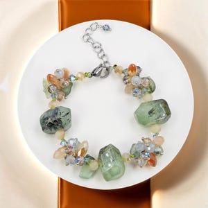 Könnte beinhalten: Ein Armband aus grünen und weißen Edelsteinen mit einem silbernen Verschluss. Das Armband verfügt über eine Vielzahl kleinerer Edelsteine in Grün-, Weiß- und Orangetönen.