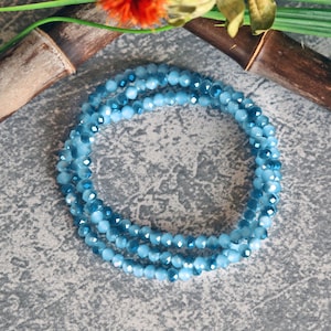 Może przedstawiać: Delikatna, warstwowa bransoletka złożona z małych, fasetowanych, jasnoniebieskich koralików. Koraliki mają subtelny połysk, tworząc urzekający efekt wizualny. Bransoletka jest prezentowana na teksturowanej, szarej powierzchni, z nutą zieleni i bambusa w tle.