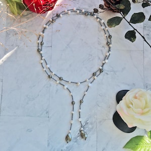 May include: A delicate necklace featuring white pearls and dark gray beads. The necklace has a Y-shaped design with two strands of pearls that meet at the top and cascade down. The necklace is adorned with two teardrop-shaped pendants.
