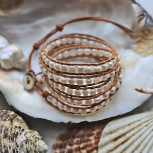 以下が含まれることがあります： 白い真珠のブレスレットが茶色の革で巻かれています。ブレスレットは白い貝殻の上に置かれています。
