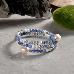 May include: A spiral bracelet featuring blue, white, and clear beads, interspersed with larger, light pink pearls. The bracelet is designed with multiple strands, creating a layered effect. The beads are small and round, with a polished finish.