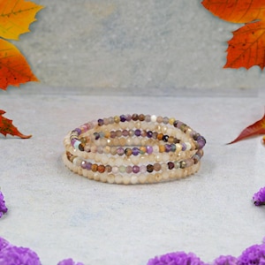 Puede incluir: Una pila de tres pulseras de cuentas. Las pulseras están hechas de pequeñas cuentas facetadas en tonos crema, marrón, morado y marrón. Las pulseras están envueltas unas alrededor de otras. El fondo es gris claro.