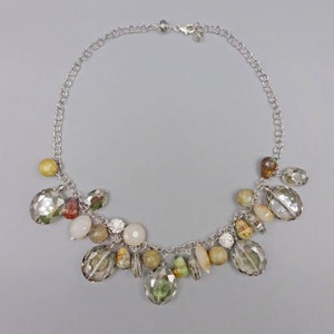 May include: Silver-toned necklace with a variety of gemstone pendants. The necklace features faceted clear, yellow, and green stones, along with smaller, colorful beads. The chain is a delicate link style, and the clasp is visible.