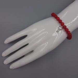 May include: Red beaded bracelet with a silver key charm. The bracelet is on a white mannequin hand.