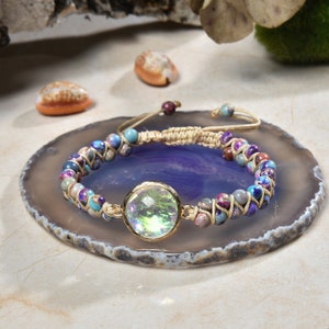 May include: A multi-coloured beaded bracelet with a gold-toned iridescent glass charm. The bracelet is made with a beige cord and features a variety of small, round beads in shades of blue, purple, and brown.