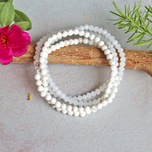May include: A white beaded bracelet with a layered design. The bracelet is made of small, round beads and is strung on a clear elastic cord.