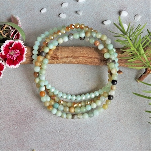 May include: Four beaded bracelets in shades of green, brown, and gold are arranged on a wooden stick. The bracelets are made of small, round beads. The background includes a gray surface, white stones, and sprigs of greenery and flowers.
