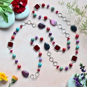 May include: A long silver chain necklace with red, purple, turquoise, and green gemstone beads. The necklace features square red gemstone accents and a large teardrop-shaped purple gemstone.