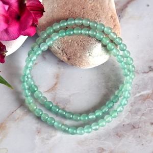 May include: A delicate, light green beaded necklace is draped over a stone. The necklace is composed of small, round beads. Pink flowers and a white bowl are in the background, set against a marble surface.