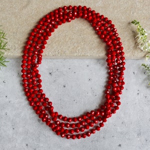 May include: A long, layered necklace composed of numerous small, faceted red beads. The necklace is arranged in a circular pattern, with multiple strands creating a full, eye-catching design. The beads have a glossy finish.