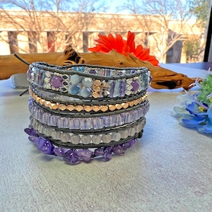 May include: A multi-layered bracelet with amethyst, gray, and clear beads. The bracelet is wrapped around a gray leather base.